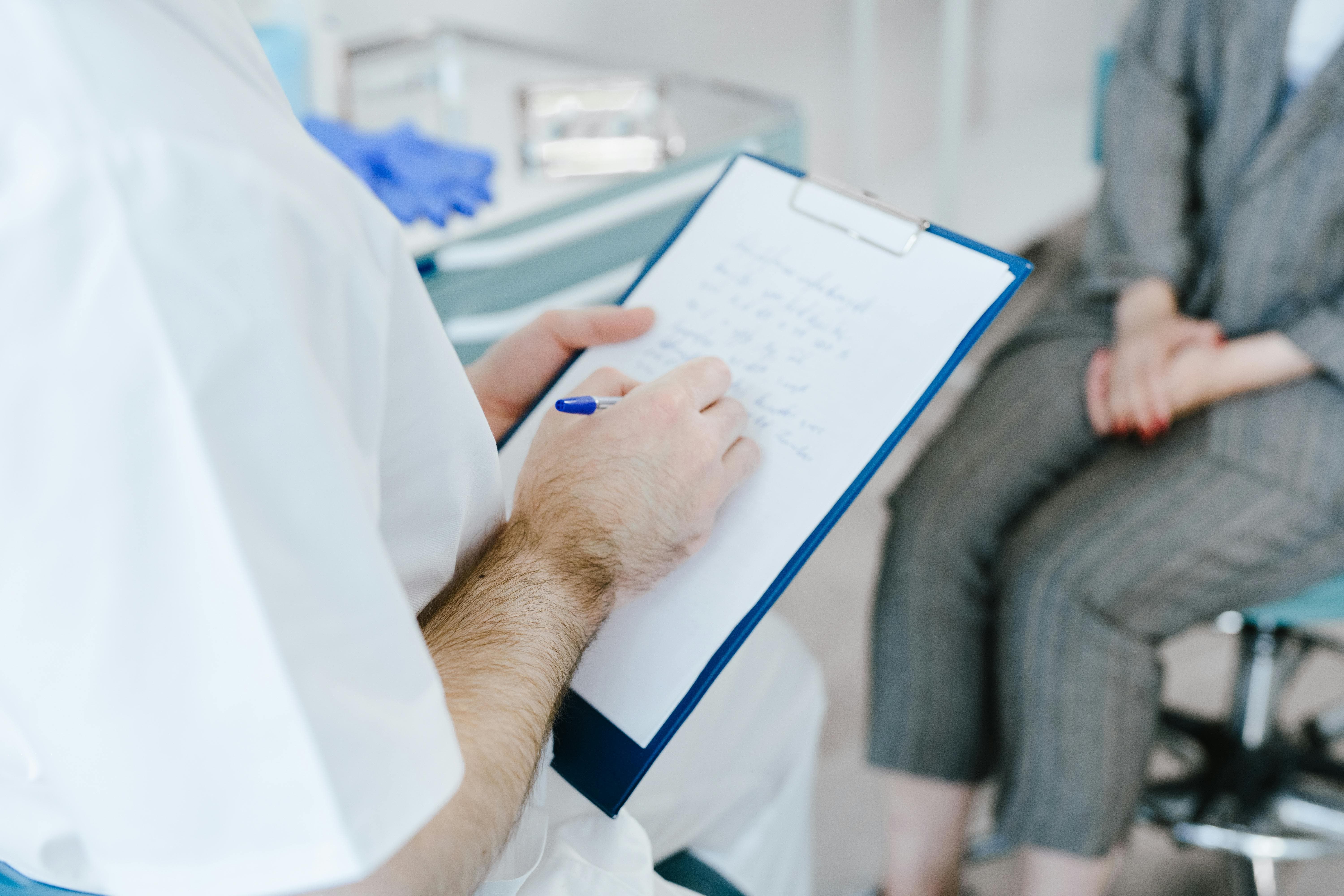 Profesional sanitario con bata blanca tomando notas en un clipboard frente a una paciente sentada en una consulta, ejemplo del sector sanitario donde las exigencias emocionales elevan el riesgo psicosocial y se exige evaluación con CoPsoQ-istas21